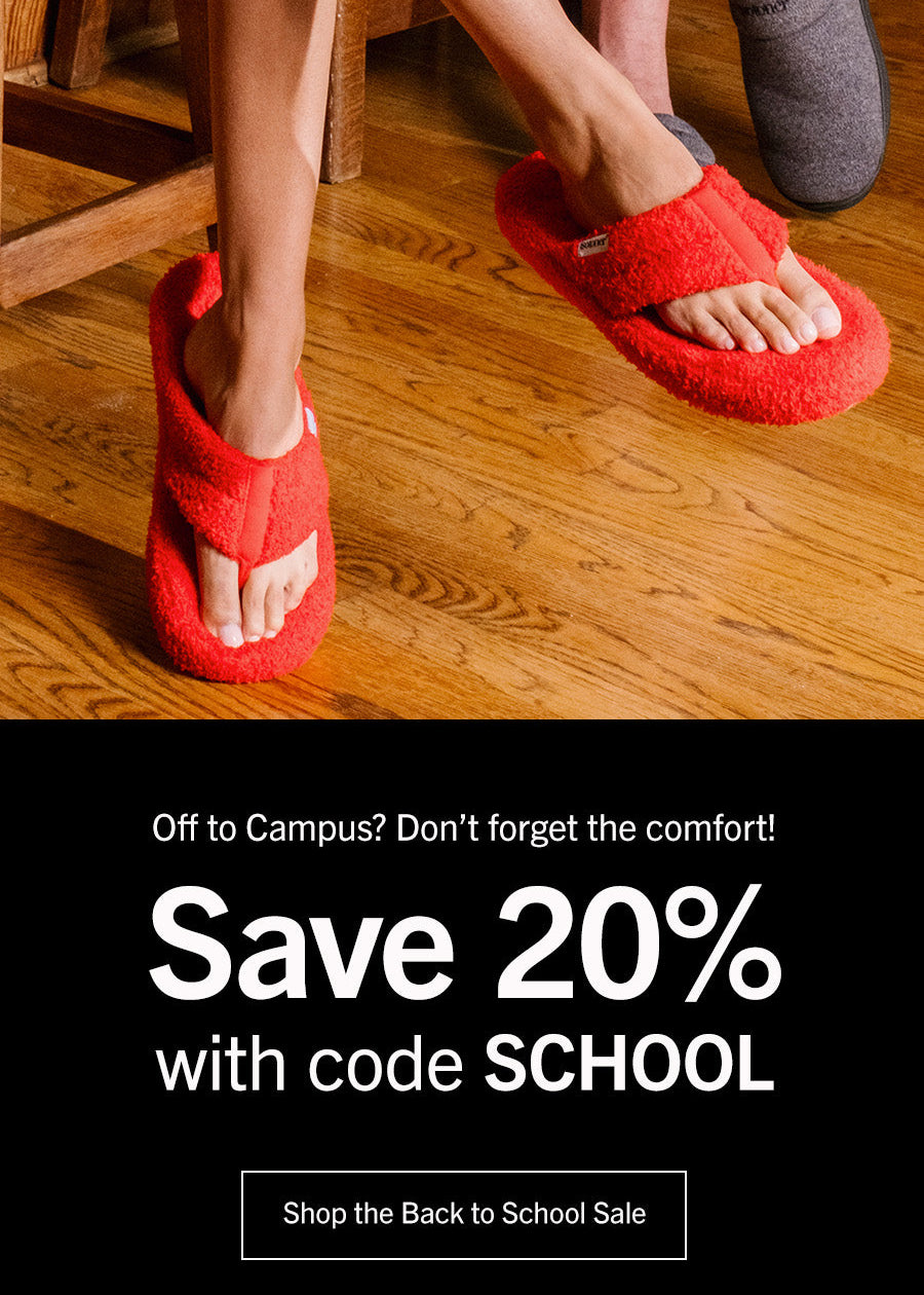A promotional lifestyle photo showing a person sitting on a wooden stool wearing bright red fuzzy slide slippers, with another person partially visible wearing gray slippers. The lower portion has a black banner with the text 'Off to Campus? Don't forget the comfort! Save 20% with code SCHOOL' and a 'Shop the Back to School Sale' button.