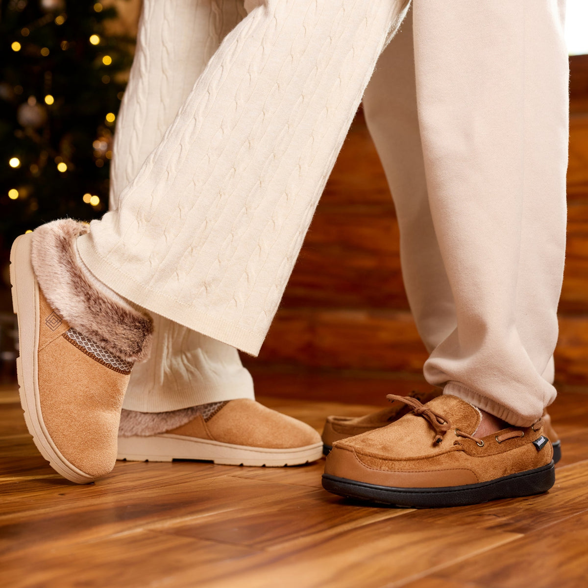 A person wearing cream knit pants and tan slip-on slippers with fuzzy lining, standing on a wooden floor in front of a decorated Christmas tree.