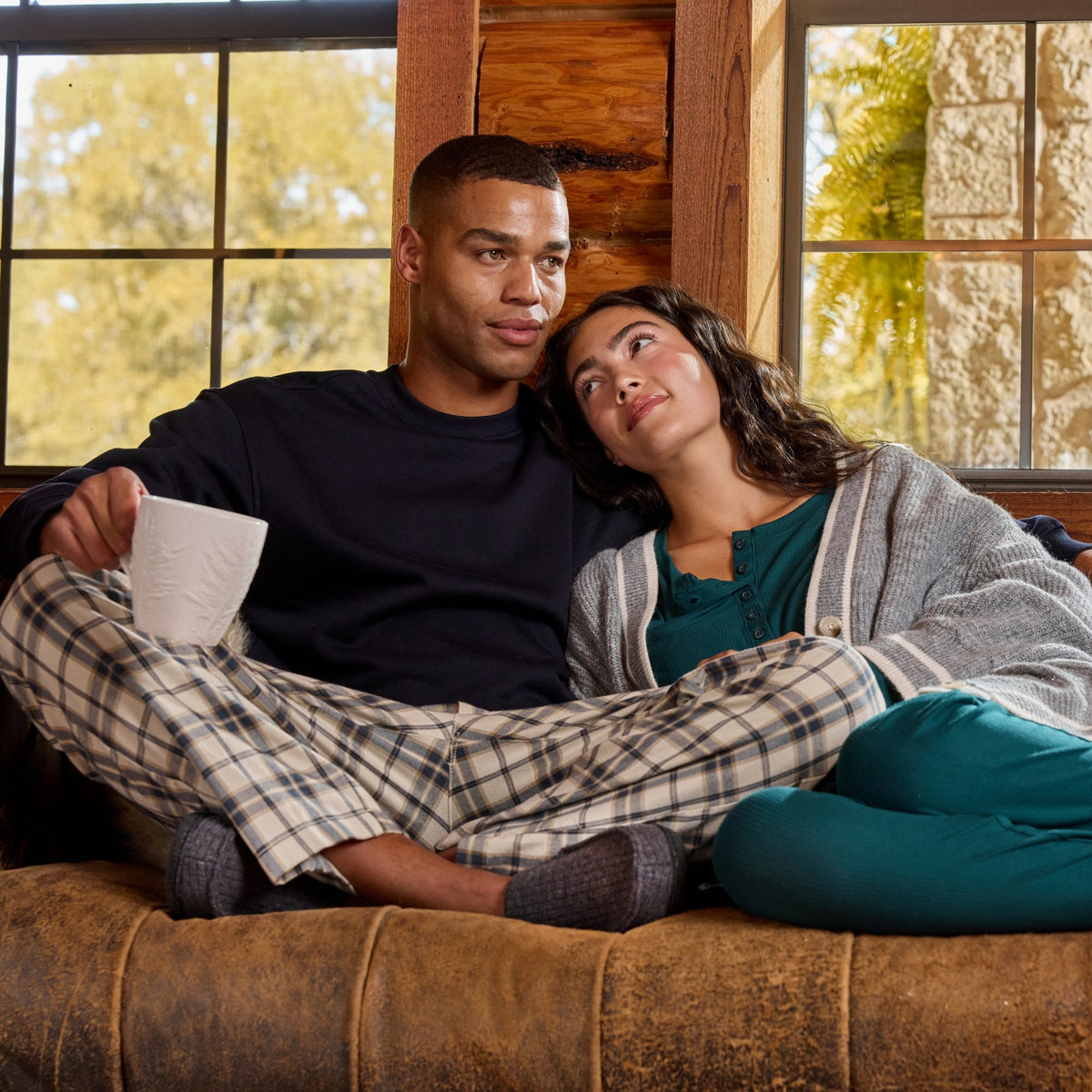 A man and a woman relax together on a brown leather couch in a rustic cabin setting. The man wears a dark navy long-sleeve shirt and plaid pajama pants, holding a white mug.