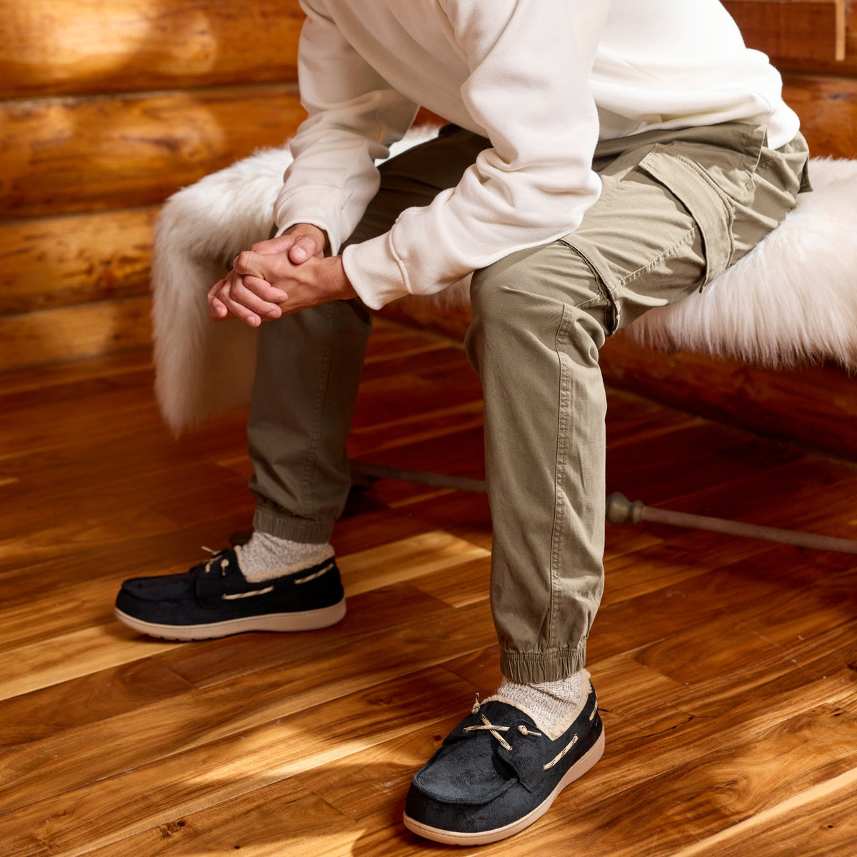 A man wearing a black beanie, cream sweatshirt, olive cargo pants, and navy boat shoes sits on a white faux-fur bench inside a rustic log cabin with a wood floor and window.