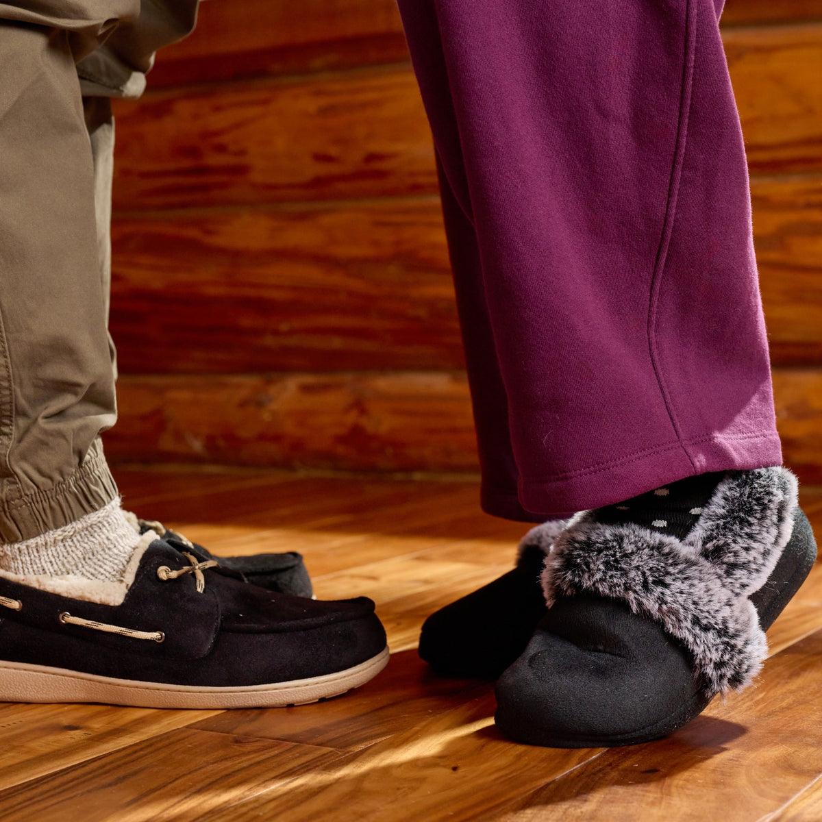 A close-up lifestyle shot of two people standing on a wooden floor in front of a log cabin wall. One person wears black boat shoes with white socks, and the other wears black slippers with gray faux-fur trim.
