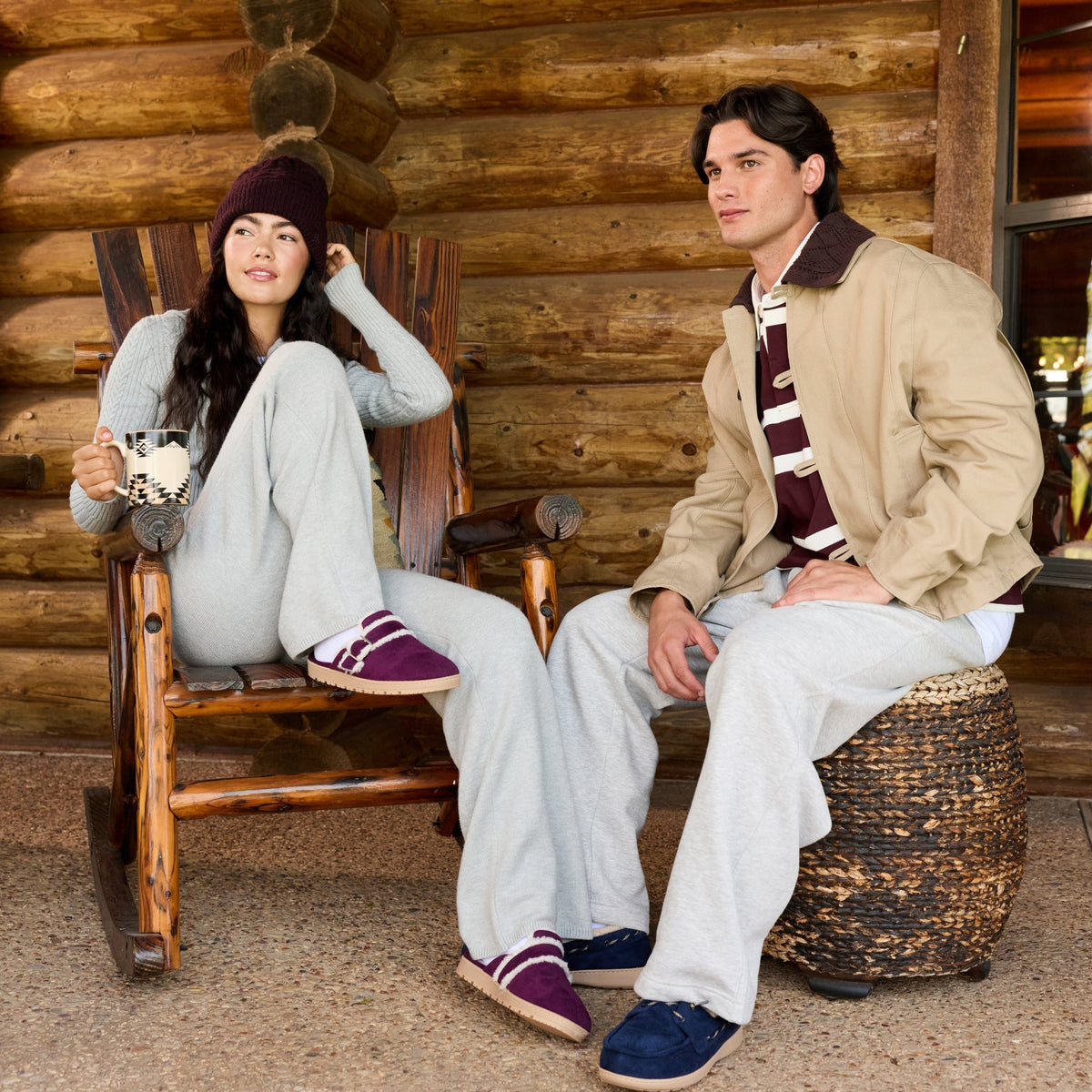 A man and a woman sit on a rustic log cabin porch. The woman wears a gray sweatshirt and sweatpants with a dark knit beanie and purple slip-on shoes, holding a mug.