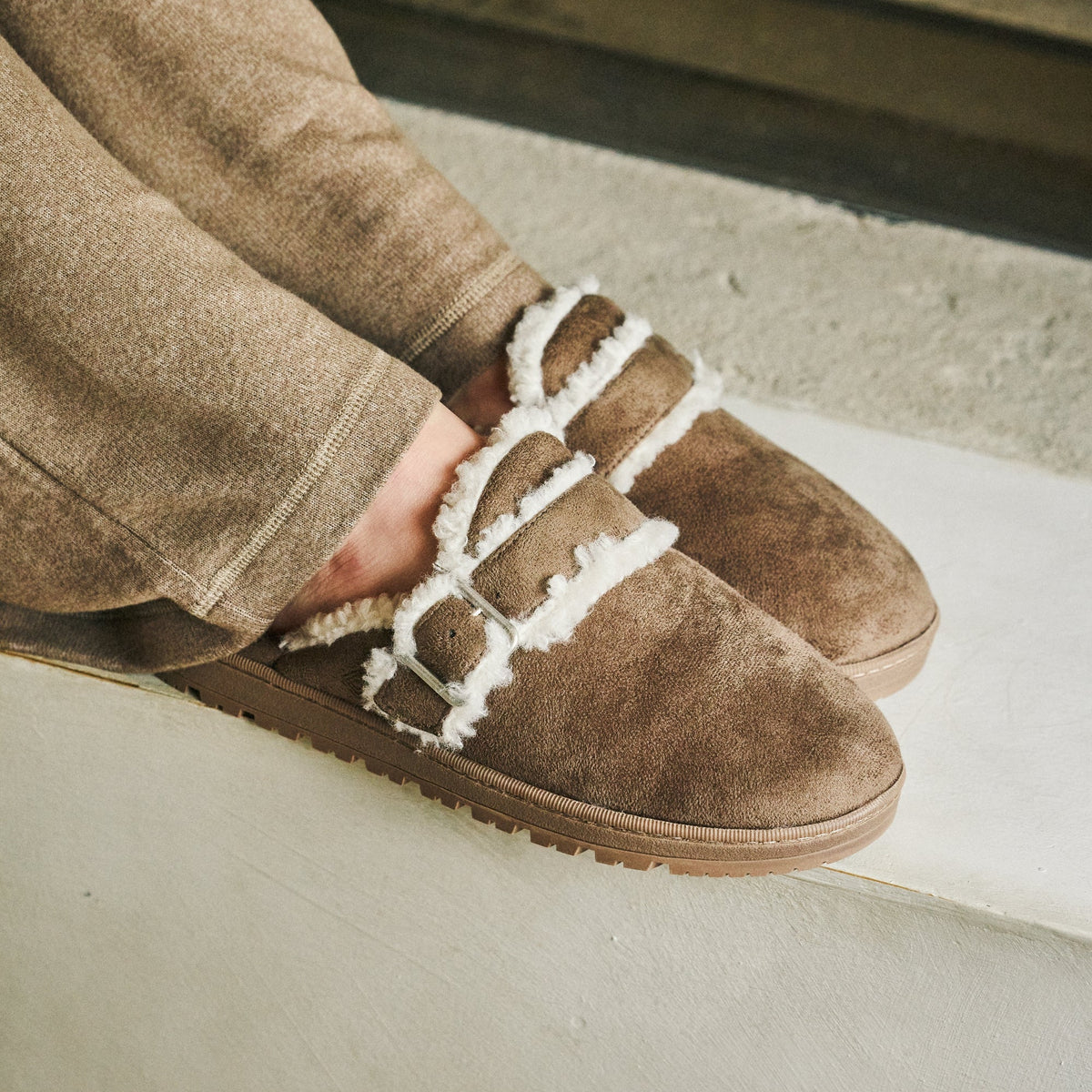 A close-up of a person wearing brown suede slip-on slippers with white faux-fur trim and decorative straps, paired with brown pants, sitting on concrete steps.