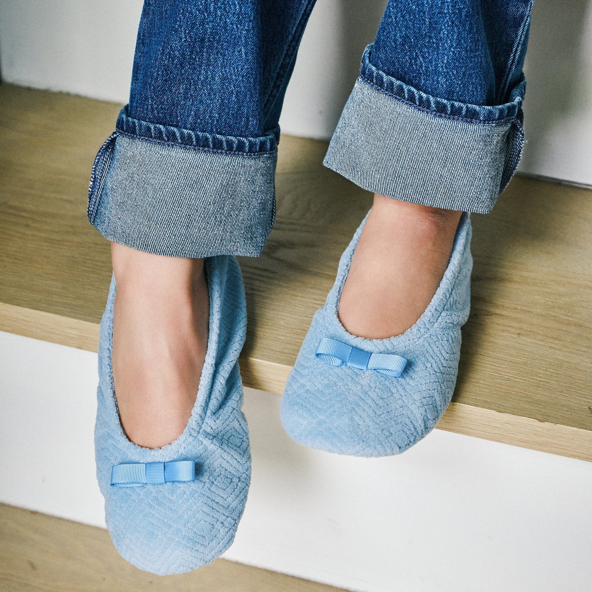 A person sitting on wooden stairs wearing light blue textured slip-on slippers with small bow details, paired with cuffed blue jeans.