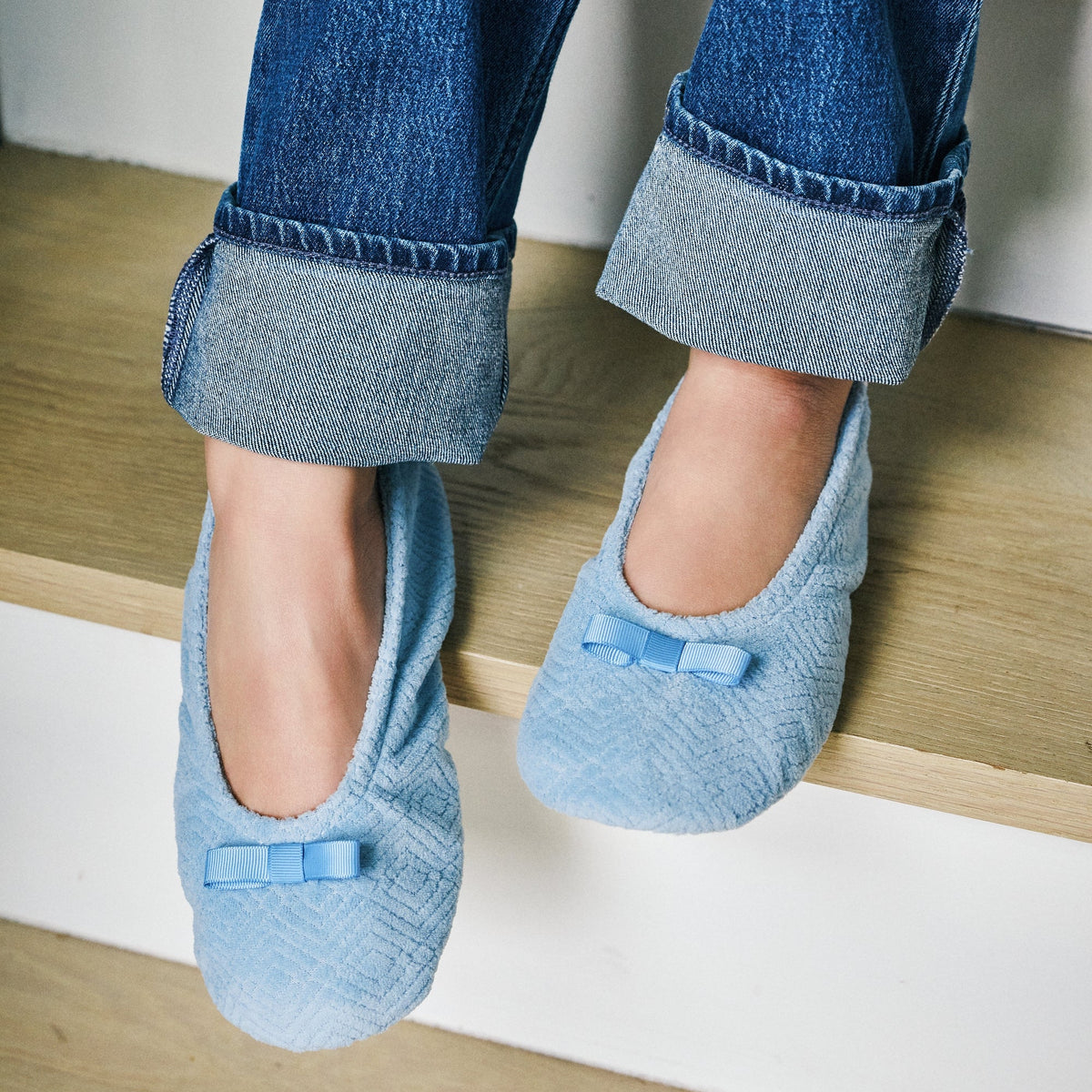 A person sitting on wooden stairs wearing light blue textured slip-on slippers with small bow details, paired with cuffed blue jeans.