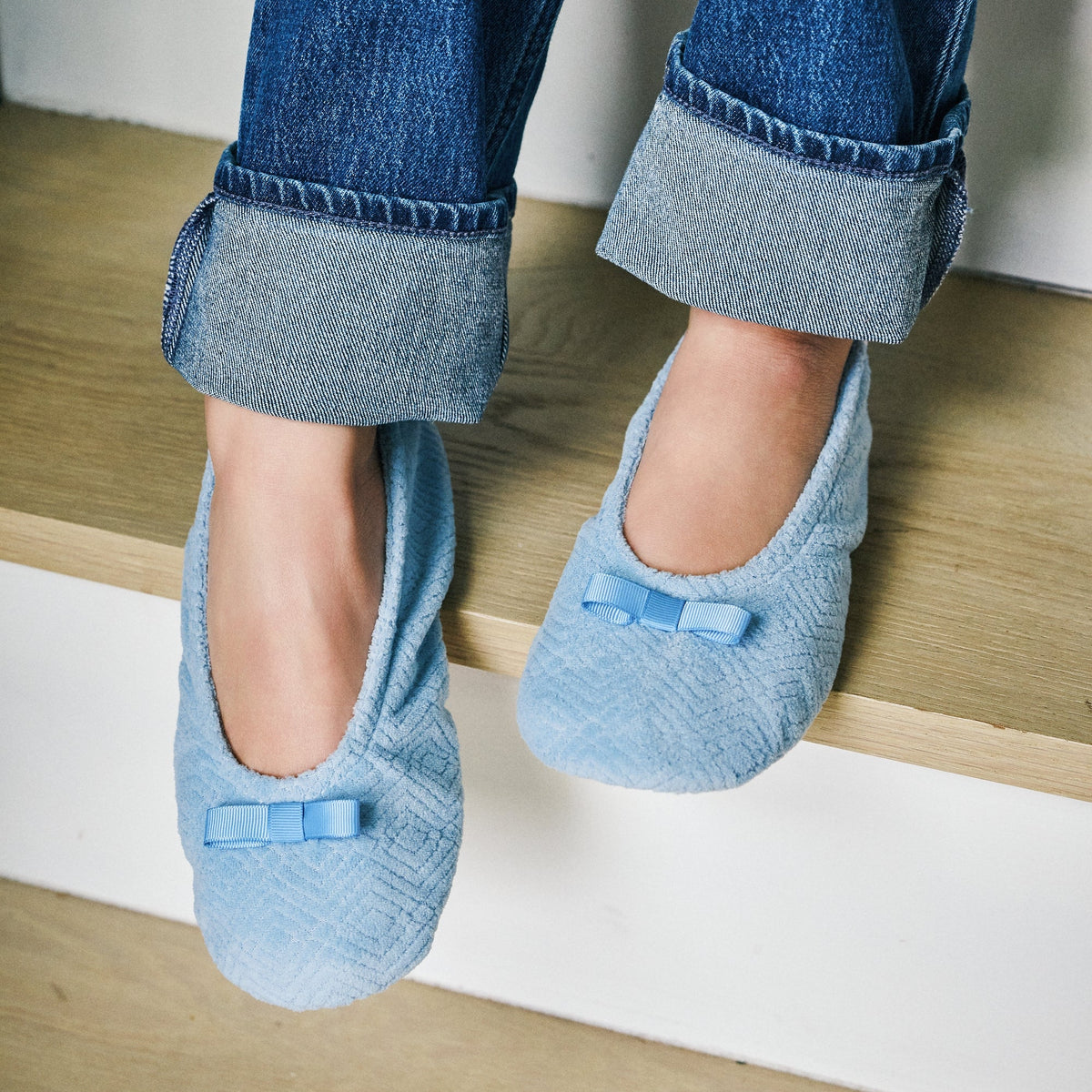 A person sitting on wooden stairs wearing light blue textured slip-on slippers with small bow details, paired with cuffed blue jeans.