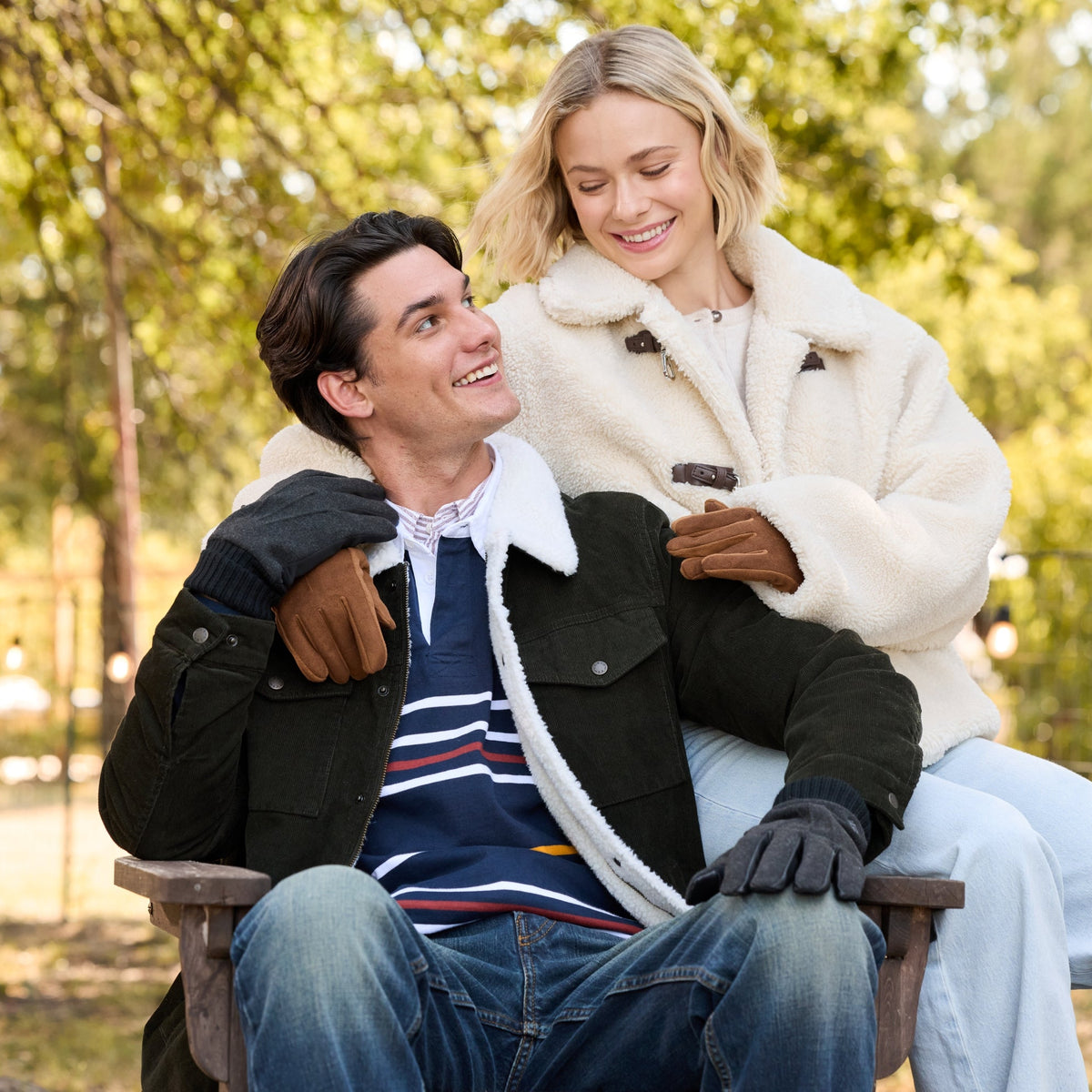 A man and a woman sitting together on a bench in an outdoor park setting. The woman is wearing a cream-colored shearling jacket and white pants, while the man wears a dark jacket over a striped sweater and jeans.

