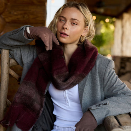 A woman wearing a gray blazer, white top, and a dark burgundy plaid scarf sits on a wooden porch next to a log cabin wall.

