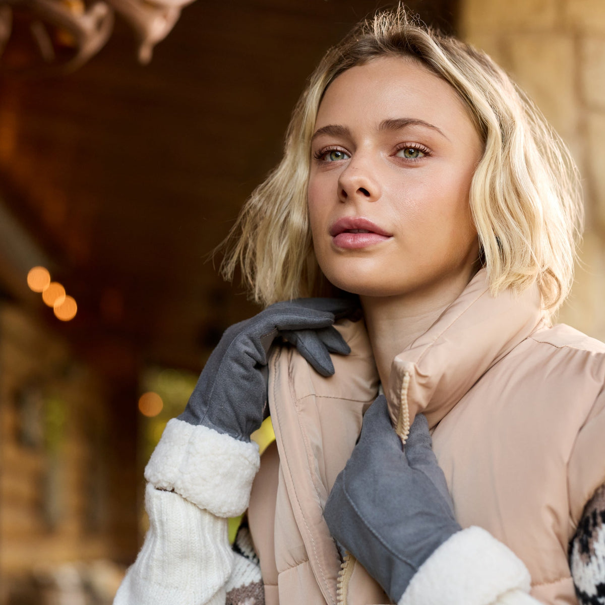 A blonde woman wearing a beige puffer vest, gray gloves, and a patterned knit sweater stands in front of a warmly lit wooden building.

