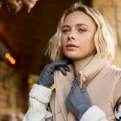 A blonde woman wearing a beige puffer vest, gray gloves, and a patterned knit sweater stands in front of a warmly lit wooden building.

