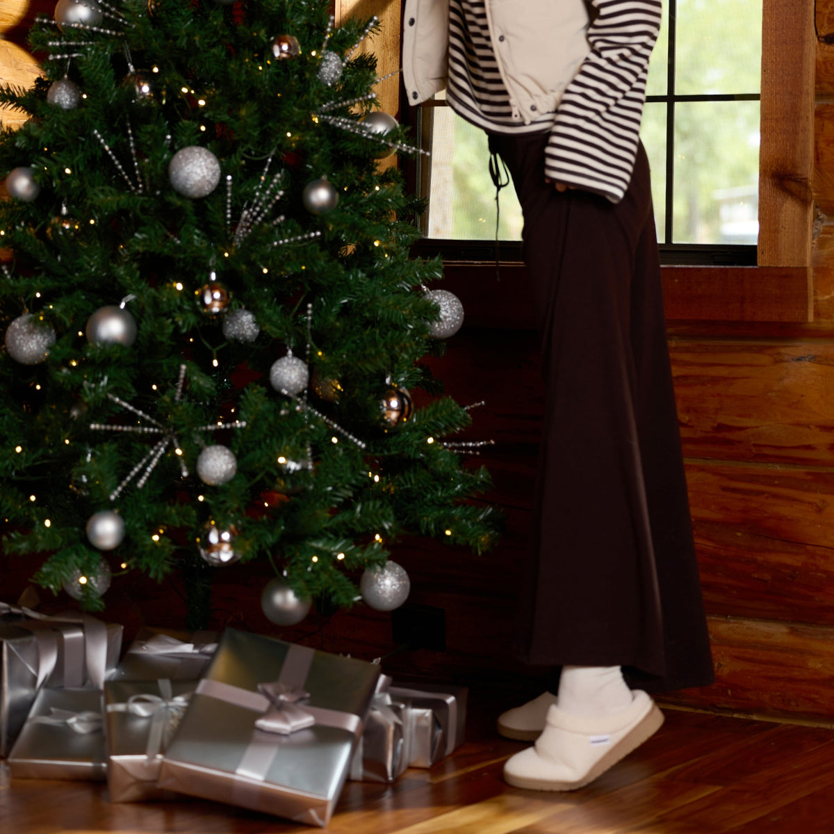 A woman in a cozy striped shirt, cream vest, dark skirt, and knit beanie is decorating a Christmas tree with silver ornaments inside a rustic wooden room. Gift boxes are wrapped at the base of the tree.


