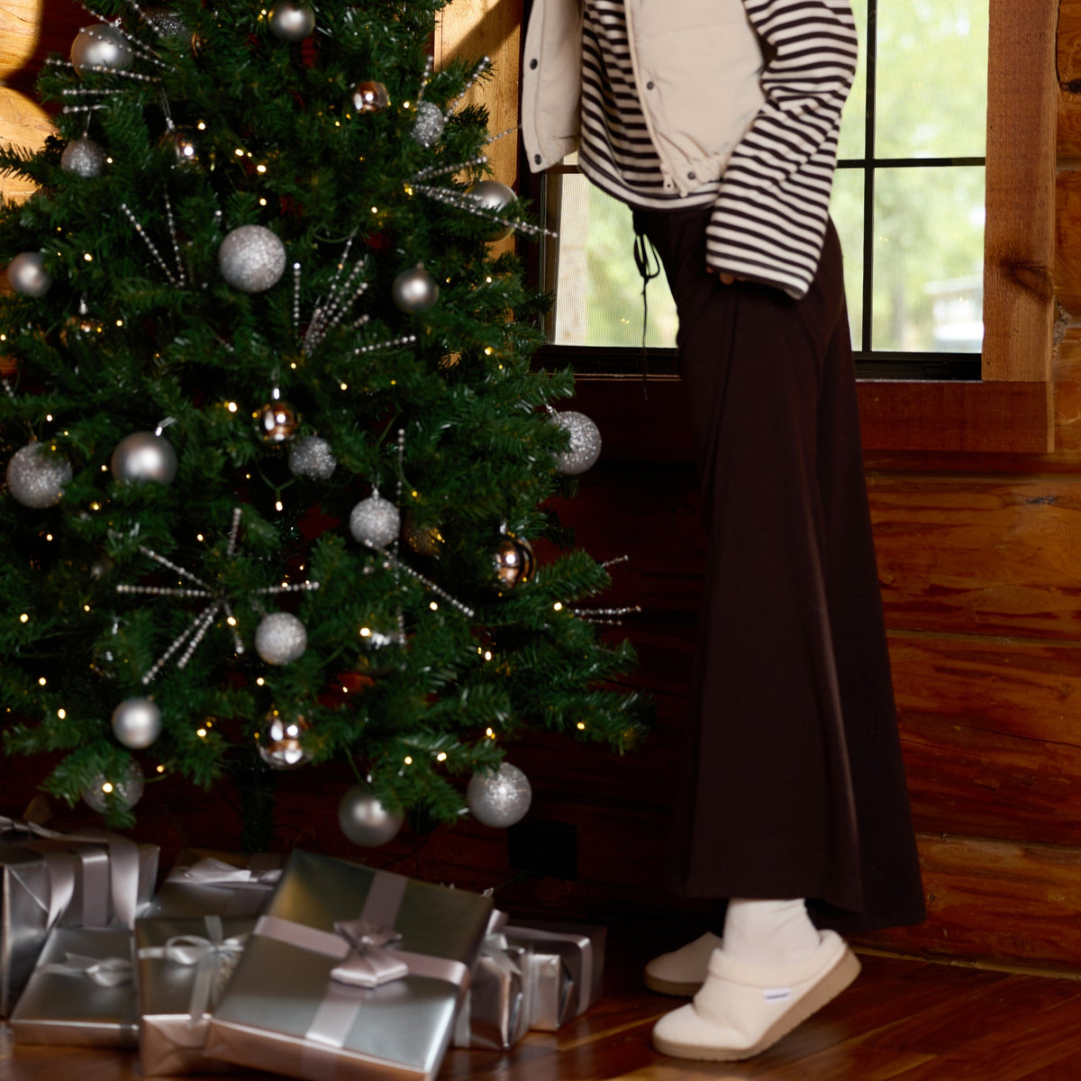 A woman in a cozy striped shirt, cream vest, dark skirt, and knit beanie is decorating a Christmas tree with silver ornaments inside a rustic wooden room. Gift boxes are wrapped at the base of the tree.

