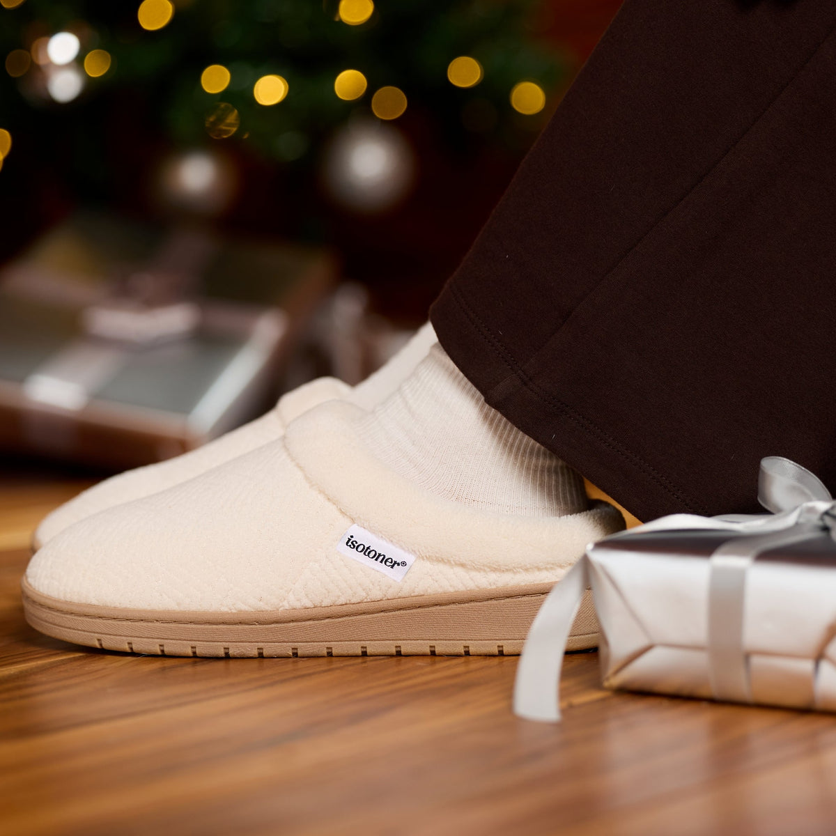 A person wearing cream-colored slip-on slippers with white socks, standing on a wooden floor next to a wrapped gift box with a silver ribbon. The background shows a blurred Christmas tree with warm lights.

