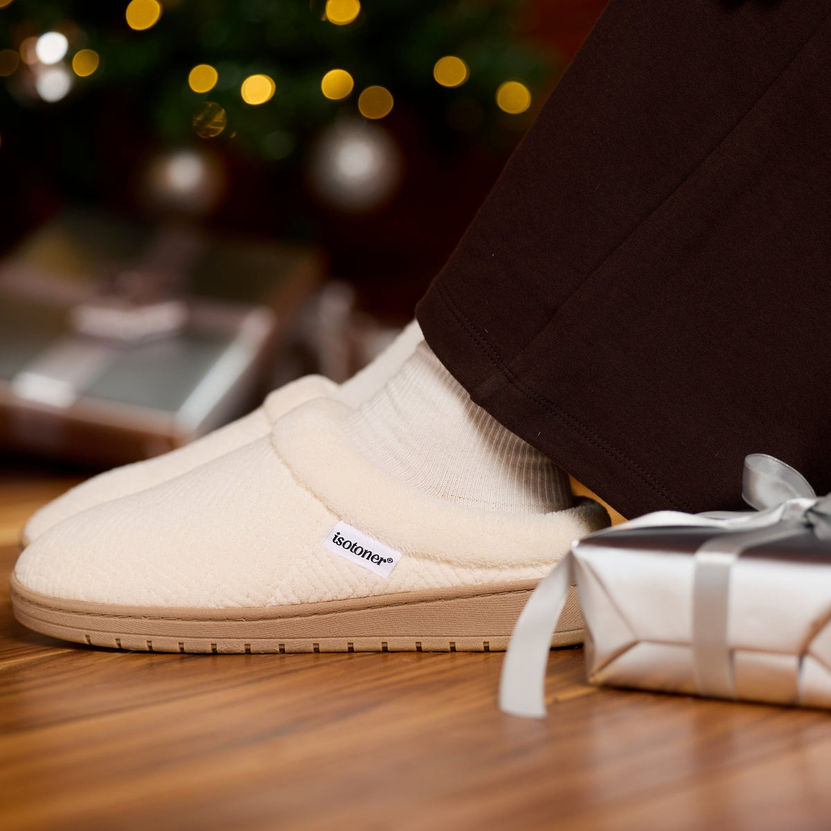 A person wearing cream-colored slip-on slippers with white socks, standing on a wooden floor next to a wrapped gift box with a silver ribbon. The background shows a blurred Christmas tree with warm lights.

