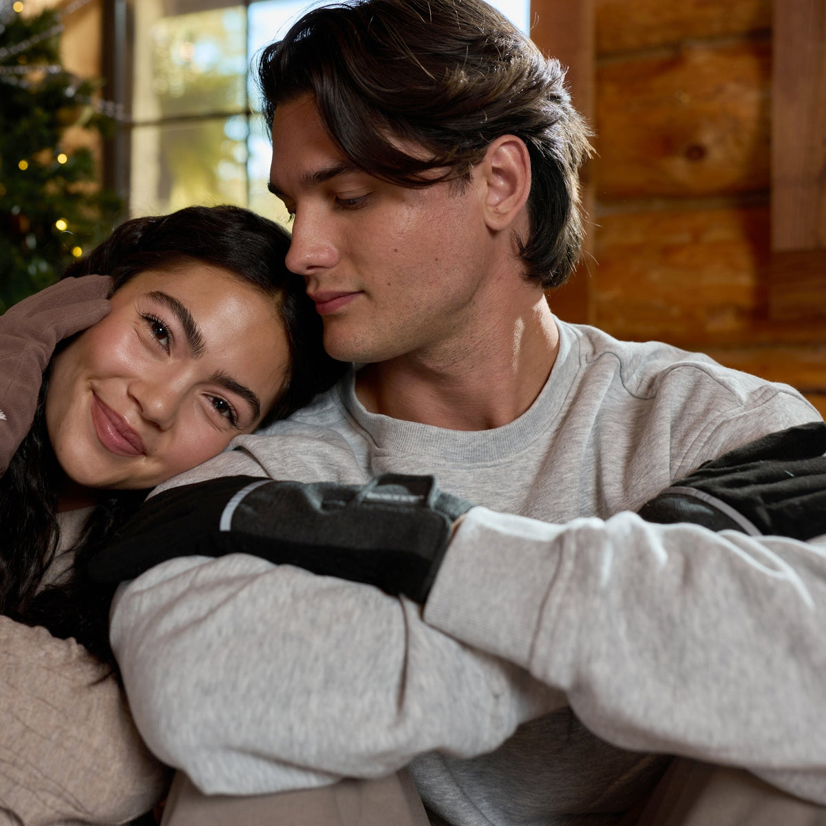 A young couple hugging indoors near a decorated Christmas tree. The woman wears a beige knit sweater and black gloves, and the man wears a light gray long-sleeve shirt and black gloves.

