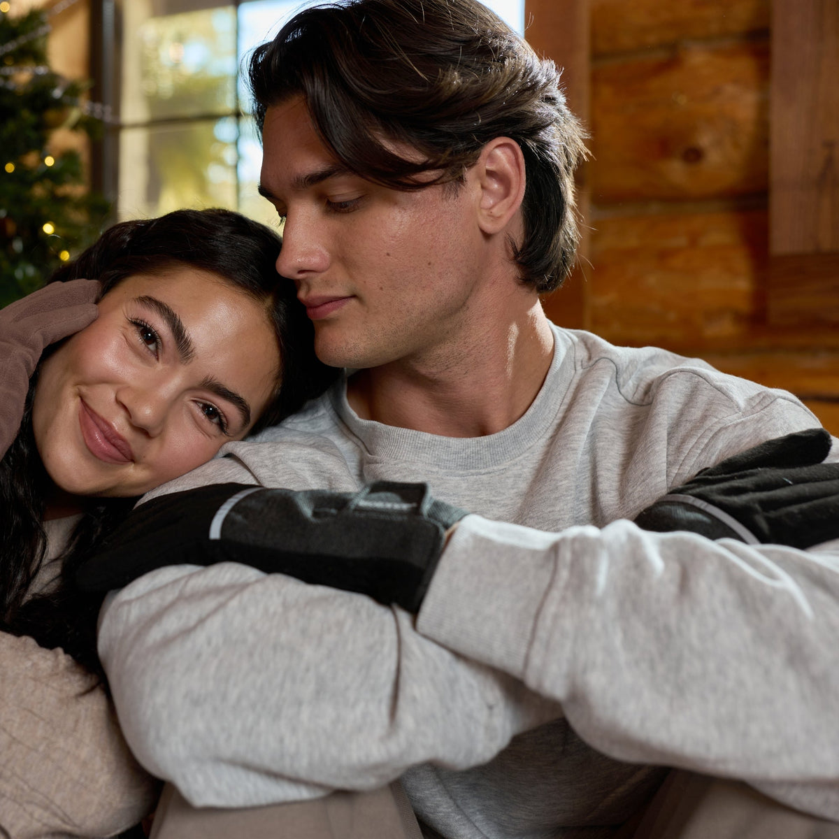 A young couple hugging indoors near a decorated Christmas tree. The woman wears a beige knit sweater and black gloves, and the man wears a light gray long-sleeve shirt and black gloves.

