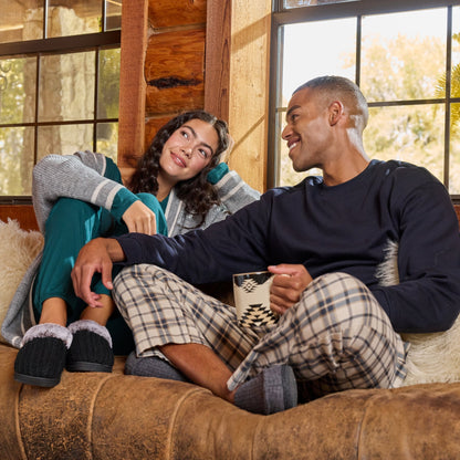 A man and a woman relax on a brown leather couch in a rustic log cabin, wearing cozy pajamas and slippers. The man wears a dark navy long-sleeve top and beige plaid pajama pants, while the woman wears a gray knit top and teal pants.

