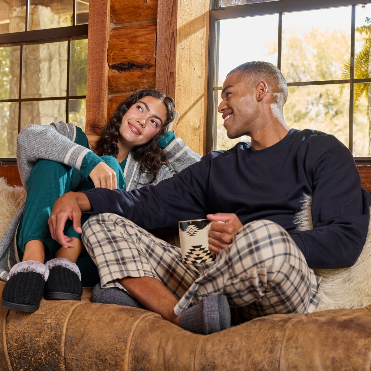 A man and a woman relax on a brown leather couch in a rustic log cabin, wearing cozy pajamas and slippers. The man wears a dark navy long-sleeve top and beige plaid pajama pants, while the woman wears a gray knit top and teal pants.

