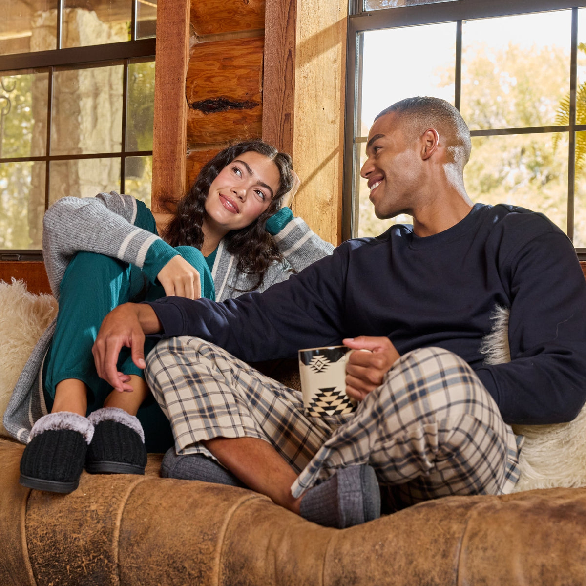 A man and a woman relax together on a brown leather couch in a rustic cabin setting. The man wears a dark navy long-sleeve shirt and plaid pajama pants, holding a white mug.

