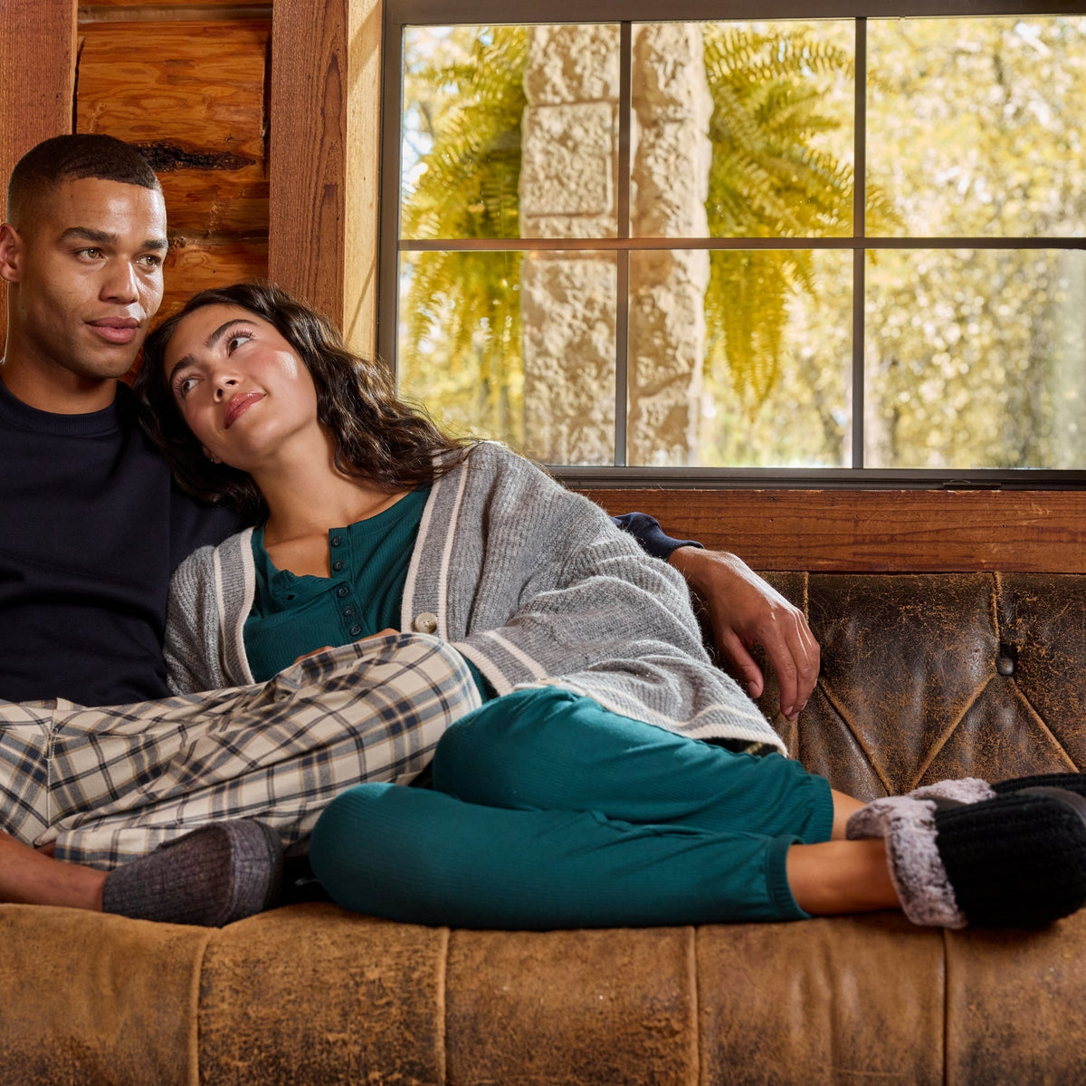 A man and a woman relax on a brown leather couch in a rustic log cabin, wearing cozy pajamas and slippers. The man wears a dark navy long-sleeve top and beige plaid pajama pants, while the woman wears a gray knit top and teal pants.

