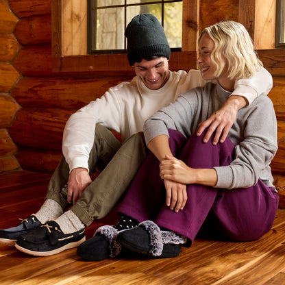 A couple sitting together on a wooden floor inside a log cabin, wearing cozy knit sweaters, pants, and casual shoes, with slippers placed in front of them.

