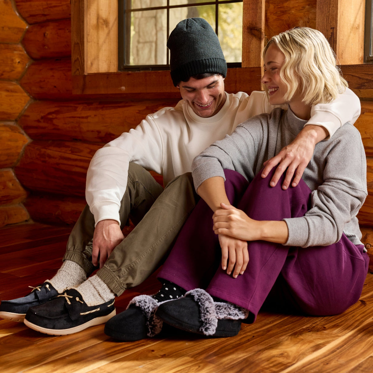 A couple sitting together on a wooden floor inside a log cabin, wearing cozy knit sweaters, pants, and casual shoes, with slippers placed in front of them.

