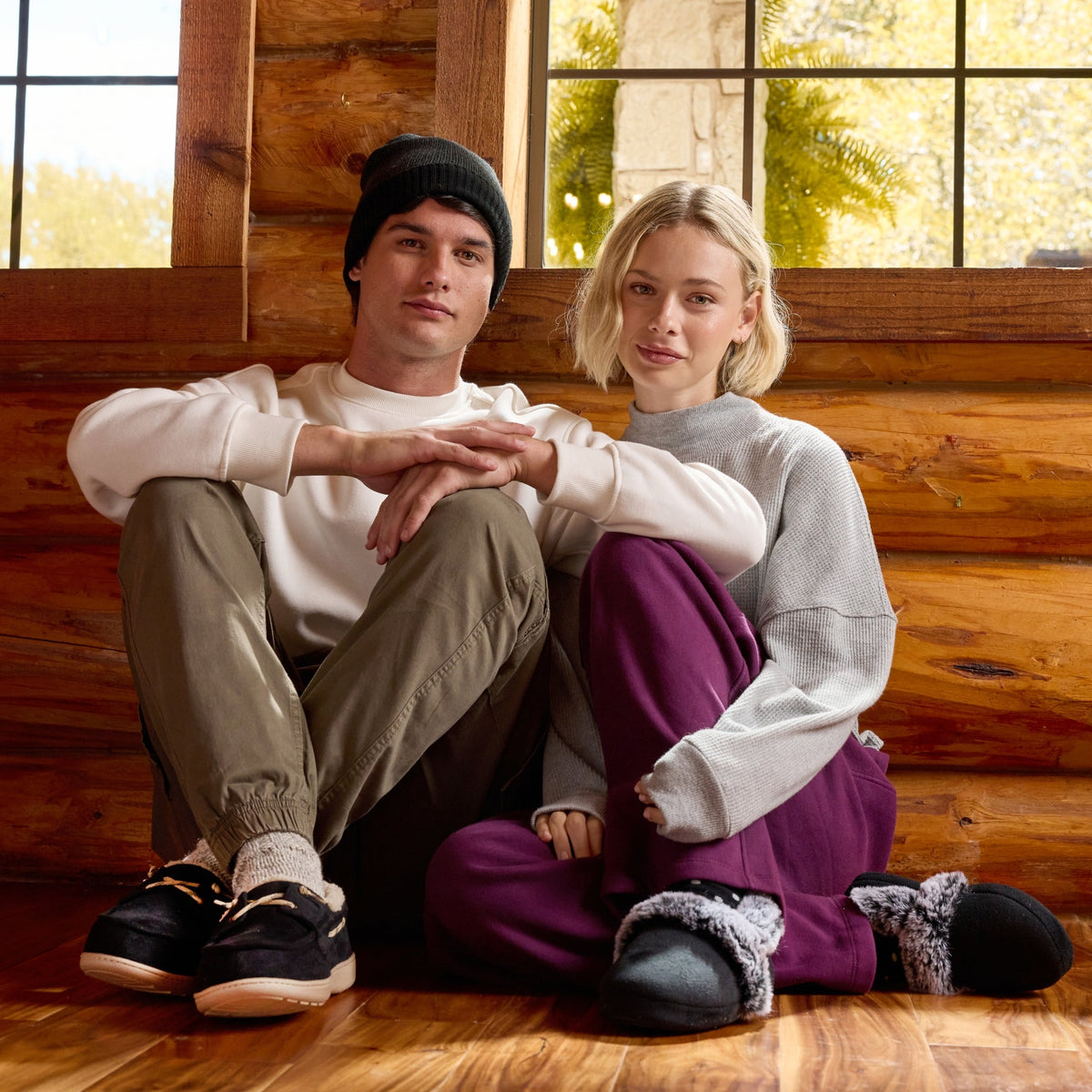 A man and a woman sitting together on a wooden floor inside a rustic log cabin, wearing cozy sweatshirts, pants, and slippers.

