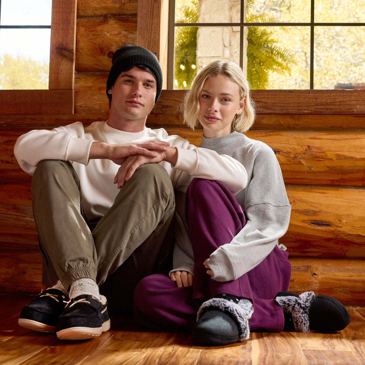 A man and a woman sitting together on a wooden floor inside a rustic log cabin, wearing cozy sweatshirts, pants, and slippers.

