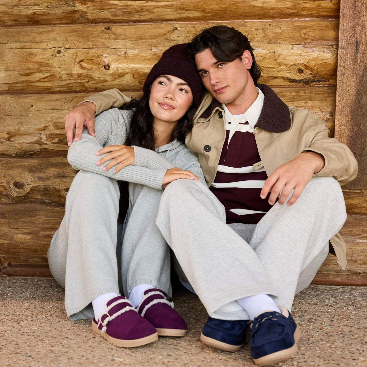 A couple sitting together in front of a rustic log cabin wall. The woman wears a light gray zip-up hoodie, light gray sweatpants, a dark maroon beanie, and purple slip-on shoes.

