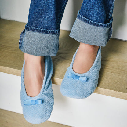 A person sitting on wooden stairs wearing light blue textured slip-on slippers with small bow details, paired with cuffed blue jeans.
