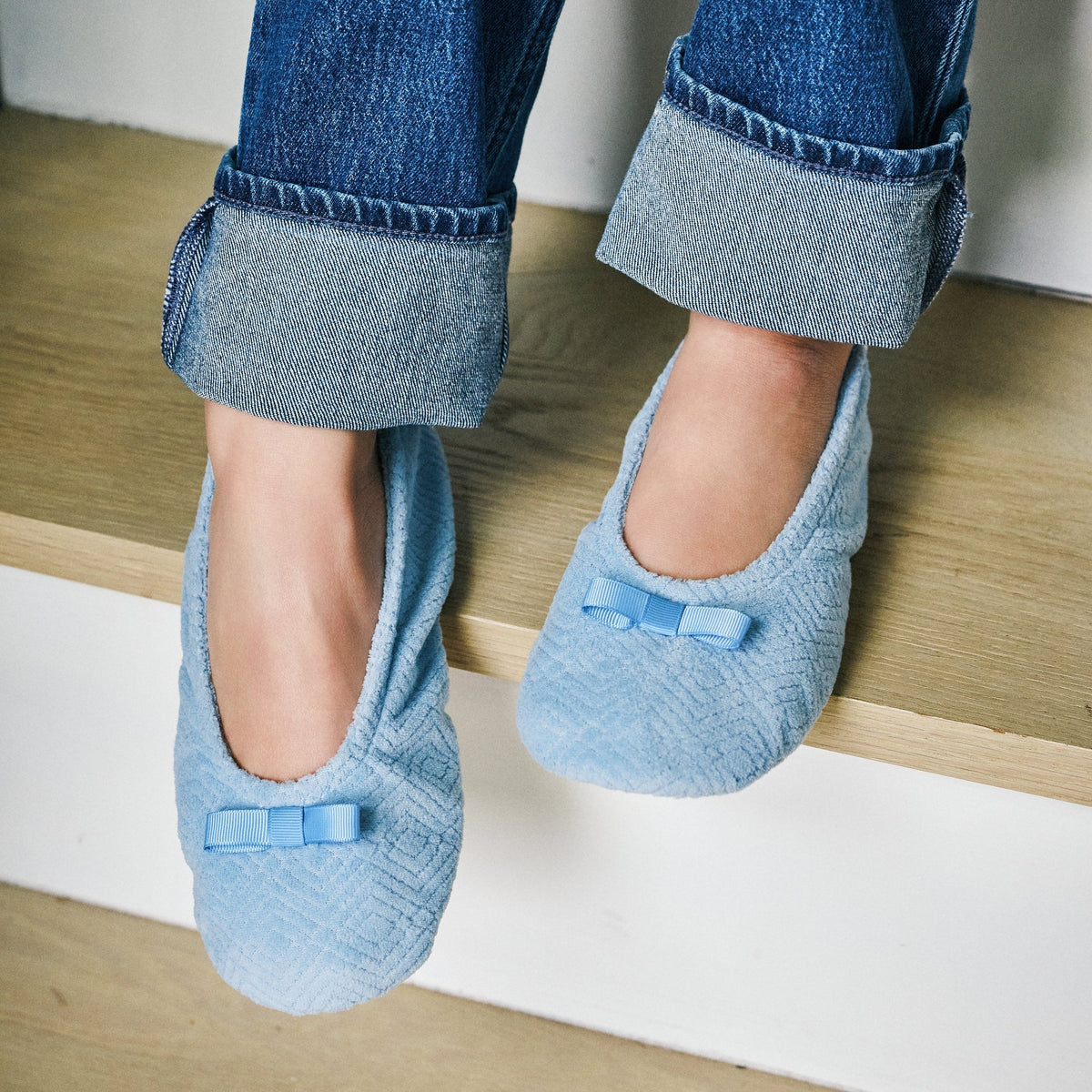 A person sitting on wooden stairs wearing light blue textured slip-on slippers with small bow details, paired with cuffed blue jeans.
