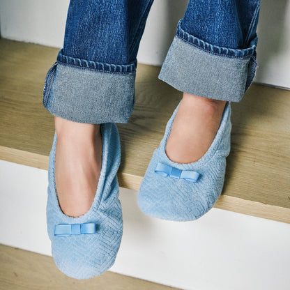 A person sitting on wooden stairs wearing light blue textured slip-on slippers with small bow details, paired with cuffed blue jeans.

