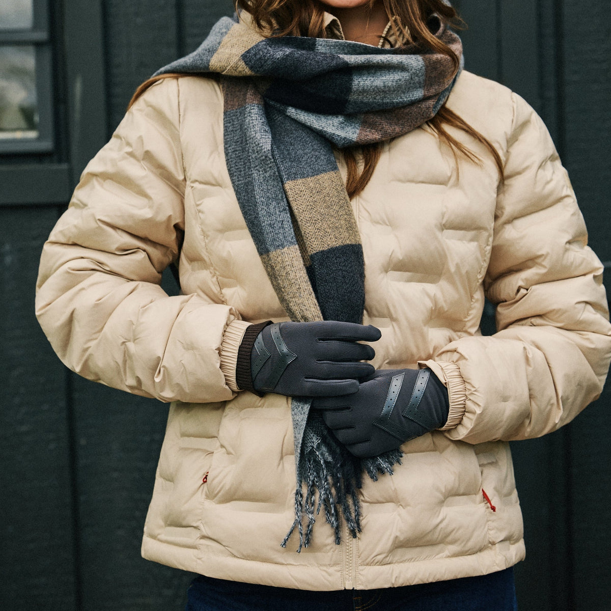 A woman wearing a beige puffer jacket, a dark gray and beige plaid scarf, black gloves, and blue jeans, standing in front of a dark gray paneled wall.

