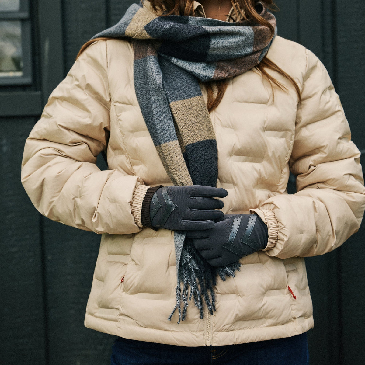 A woman wearing a beige puffer jacket, a dark gray and beige plaid scarf, black gloves, and blue jeans, standing in front of a dark gray paneled wall.

