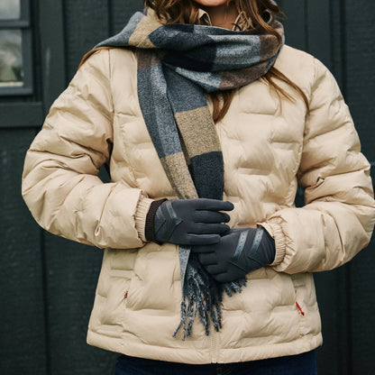 A woman wearing a beige puffer jacket, a dark gray and beige plaid scarf, black gloves, and blue jeans, standing in front of a dark gray paneled wall.


