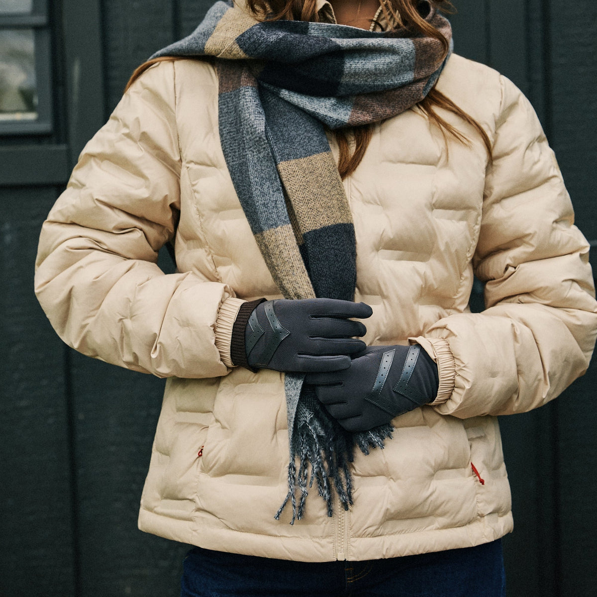 A woman wearing a beige puffer jacket, a dark gray and beige plaid scarf, black gloves, and blue jeans, standing in front of a dark gray paneled wall.

