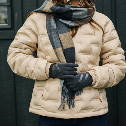 A woman wearing a beige puffer jacket, a dark gray and beige plaid scarf, black gloves, and blue jeans, standing in front of a dark gray paneled wall.

