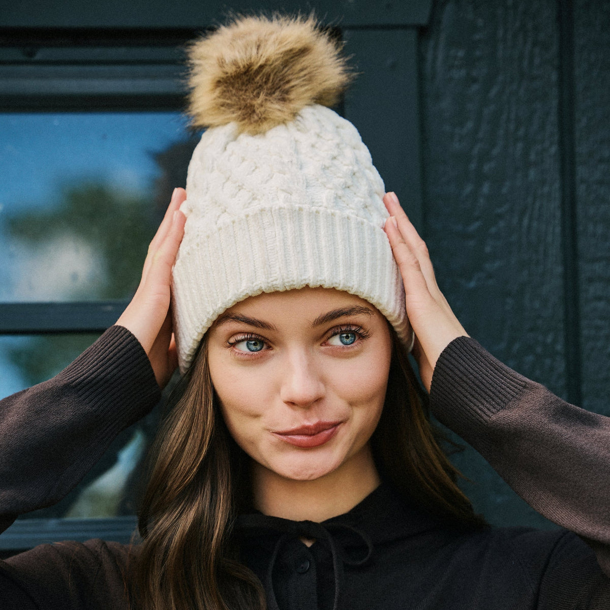 A woman wearing a cream cable-knit beanie with a brown faux-fur pom-pom, posing with her hands on her head against a dark wooden door background.

