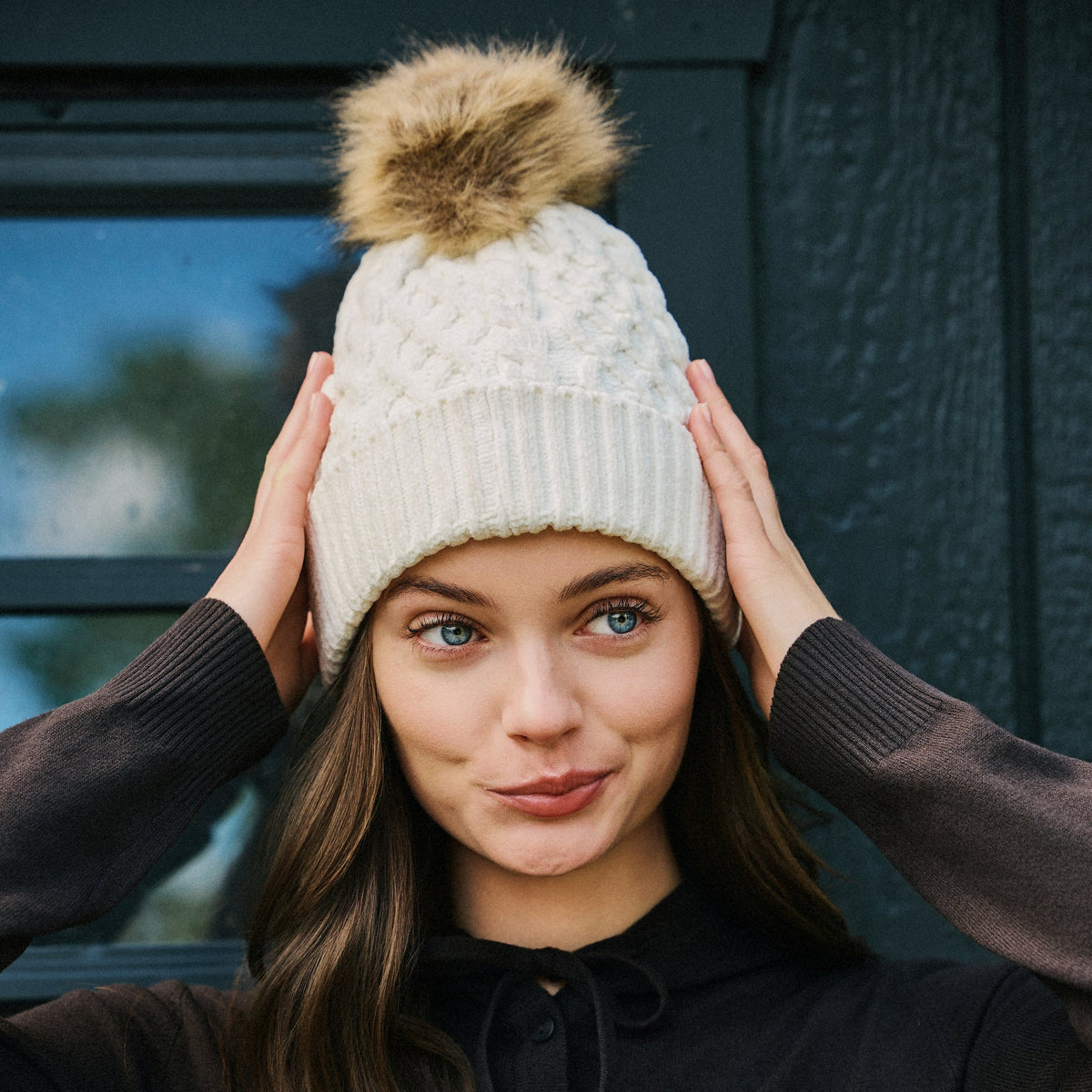 A woman wearing a cream cable-knit beanie with a brown faux-fur pom-pom, posing with her hands on her head against a dark wooden door background.
