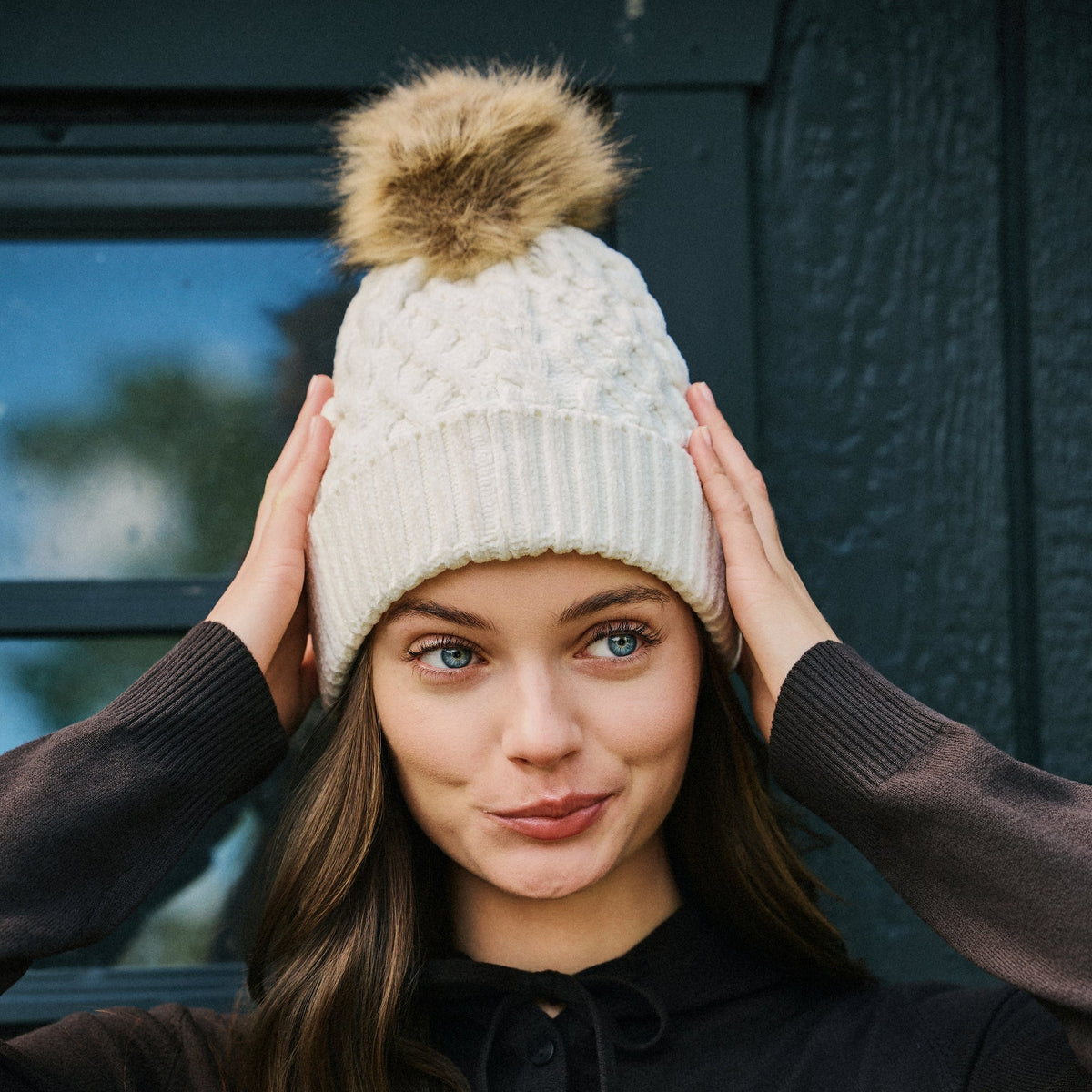 A woman wearing a cream cable-knit beanie with a brown faux-fur pom-pom, posing with her hands on her head against a dark wooden door background.


