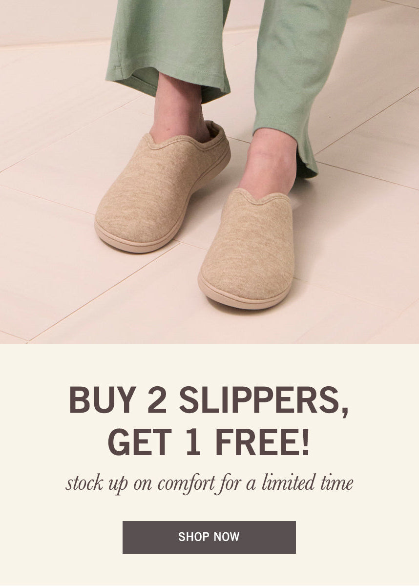 A person wearing beige slip-on slippers with sage green pants, standing on a light beige tiled floor. The lower half of the image features a promotional banner with the text 'BUY 2 SLIPPERS, GET 1 FREE!' and a 'SHOP NOW' button.