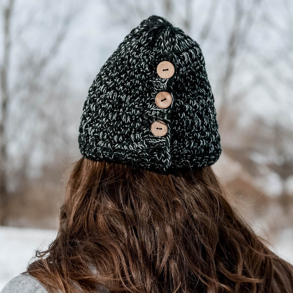 Women’s Chunky Button Hat in Black and Grey on figure. Model facing a wooded area in the snow showing the buttons on the hat