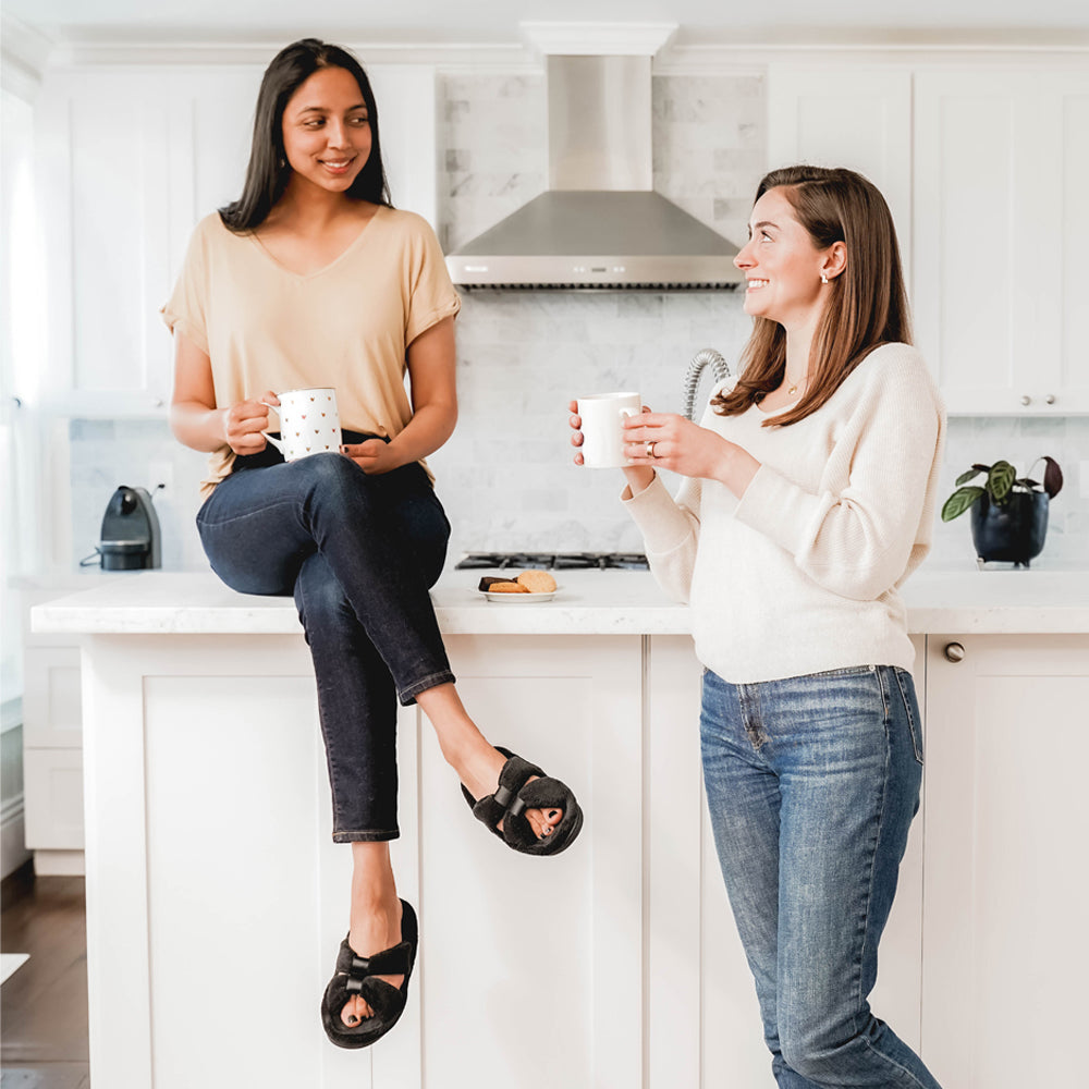 Signature Women’s Microterry with Satin X-Slide Slippers in black on figure. Two female models talking over coffee. One sitting on the kitchen counter wearing her black x-slides.