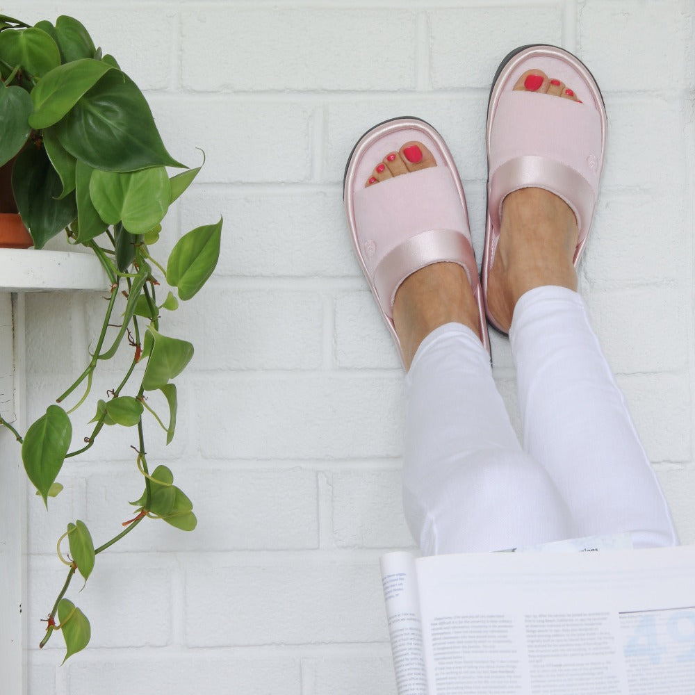 womens microterry satin trim wider width slipper in pink on figure. Model has her feet up on a brick wall with a house plant beside her and a magazine in her hands